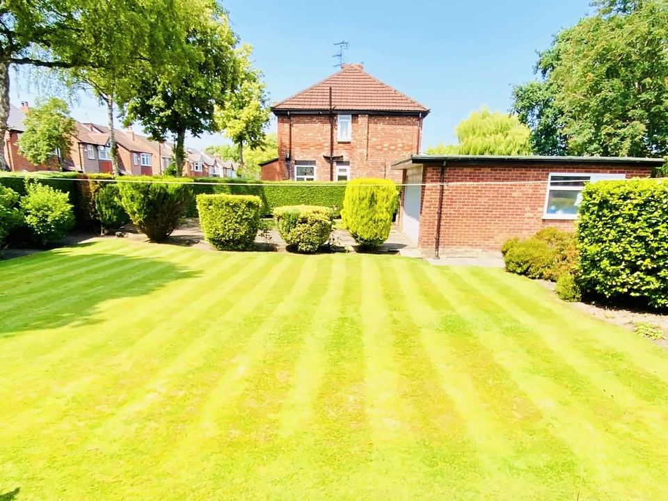 Striped lawn in front of clipped shrubs and a hedge, with a brick garage and house behind