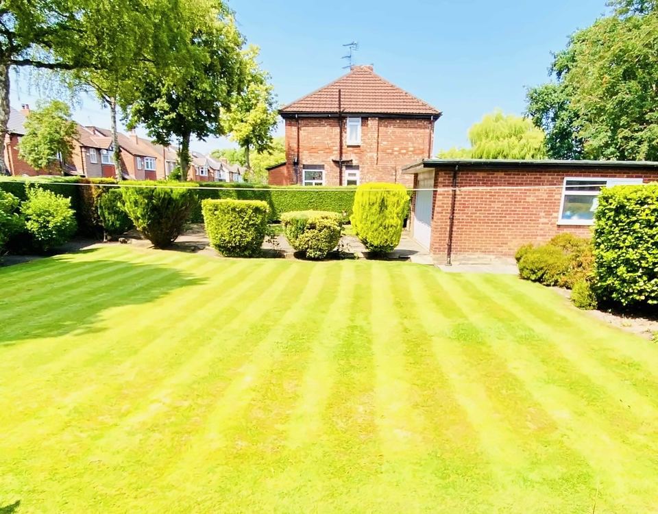 Striped lawn in front of clipped shrubs and a hedge, with a brick garage and house behind